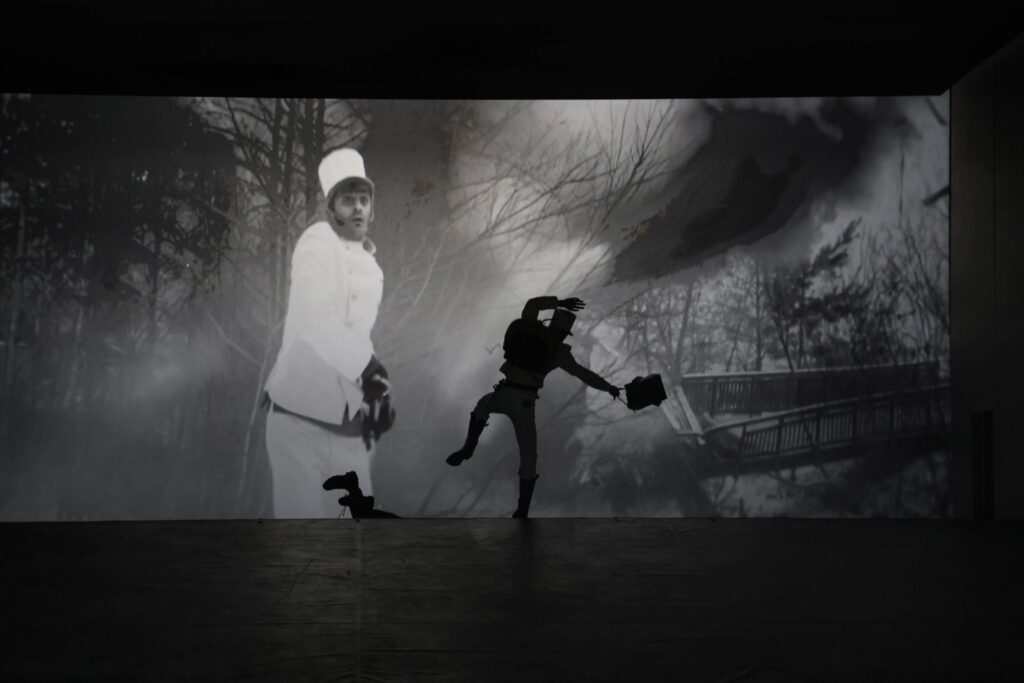 Eine schwarze Gestalt steht vor einer Videowand.